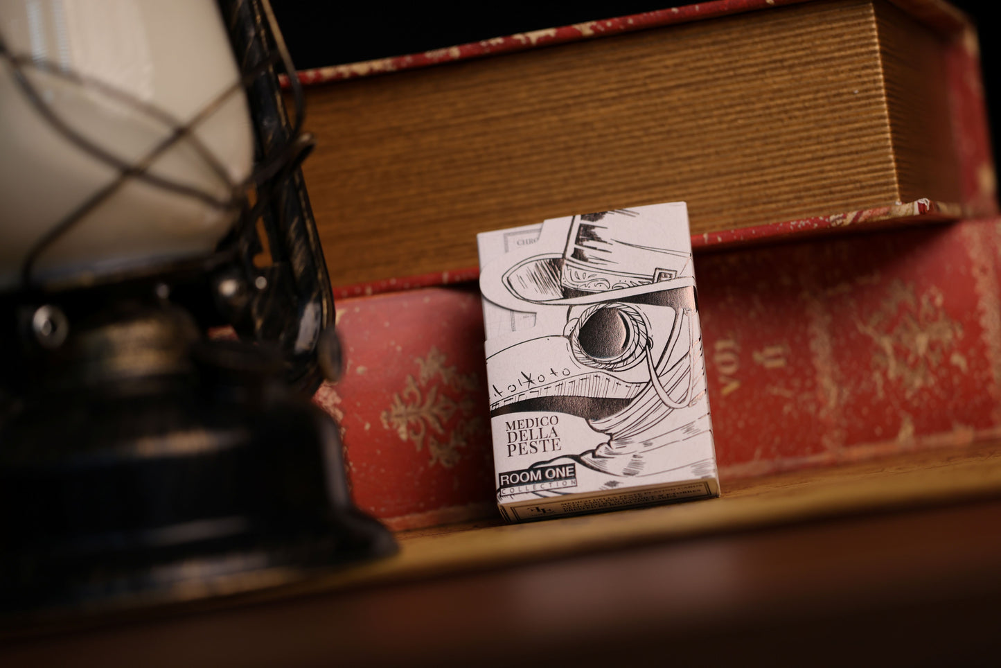 A deck of Plague Doctor playing cards with a plague doctor's mask design on the back, placed on a surface next to an old-fashioned lamp and a book.