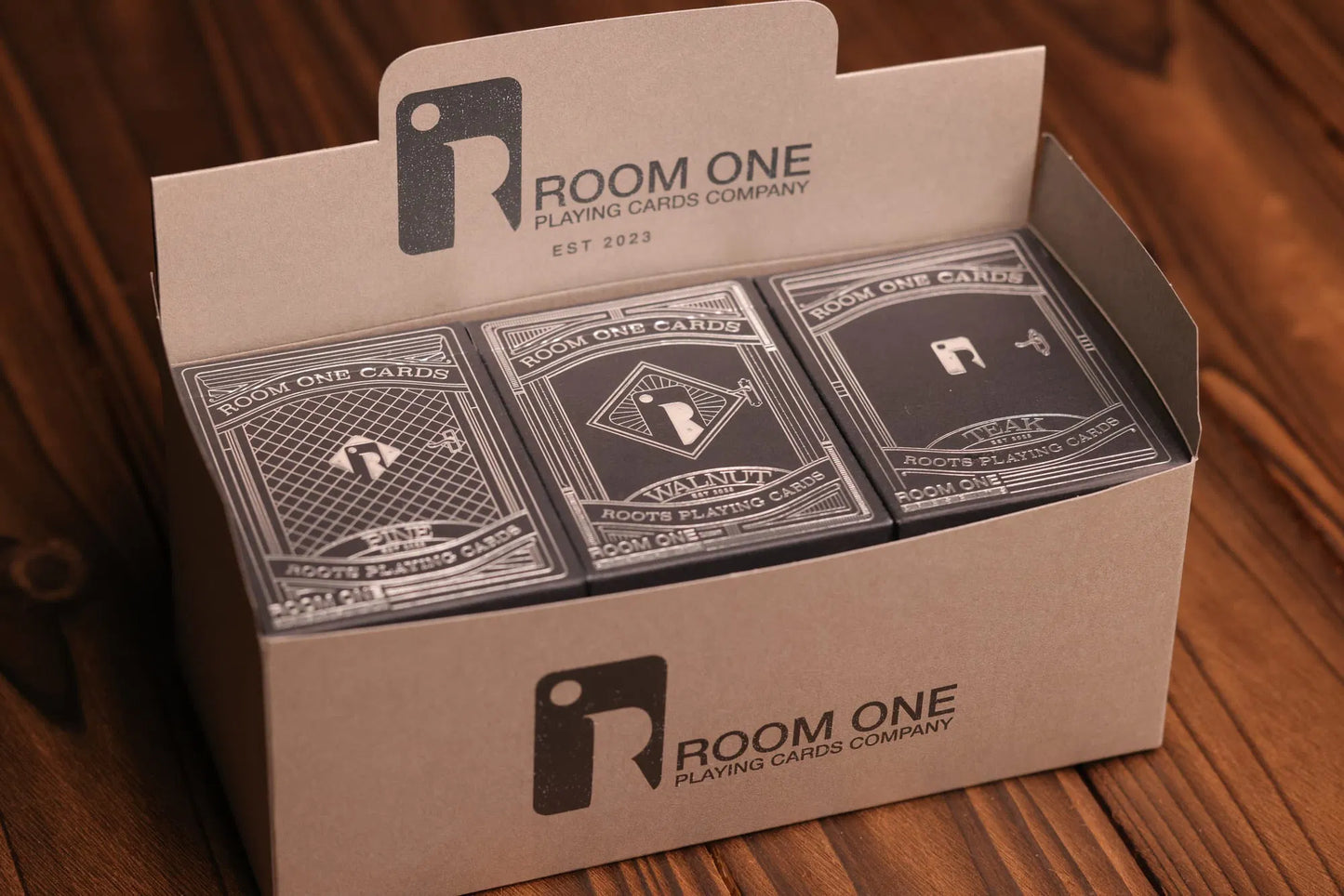 A box containing 12 decks of Roots playing cards in columns of 3, with each column displaying a different deck, placed on a wooden surface.