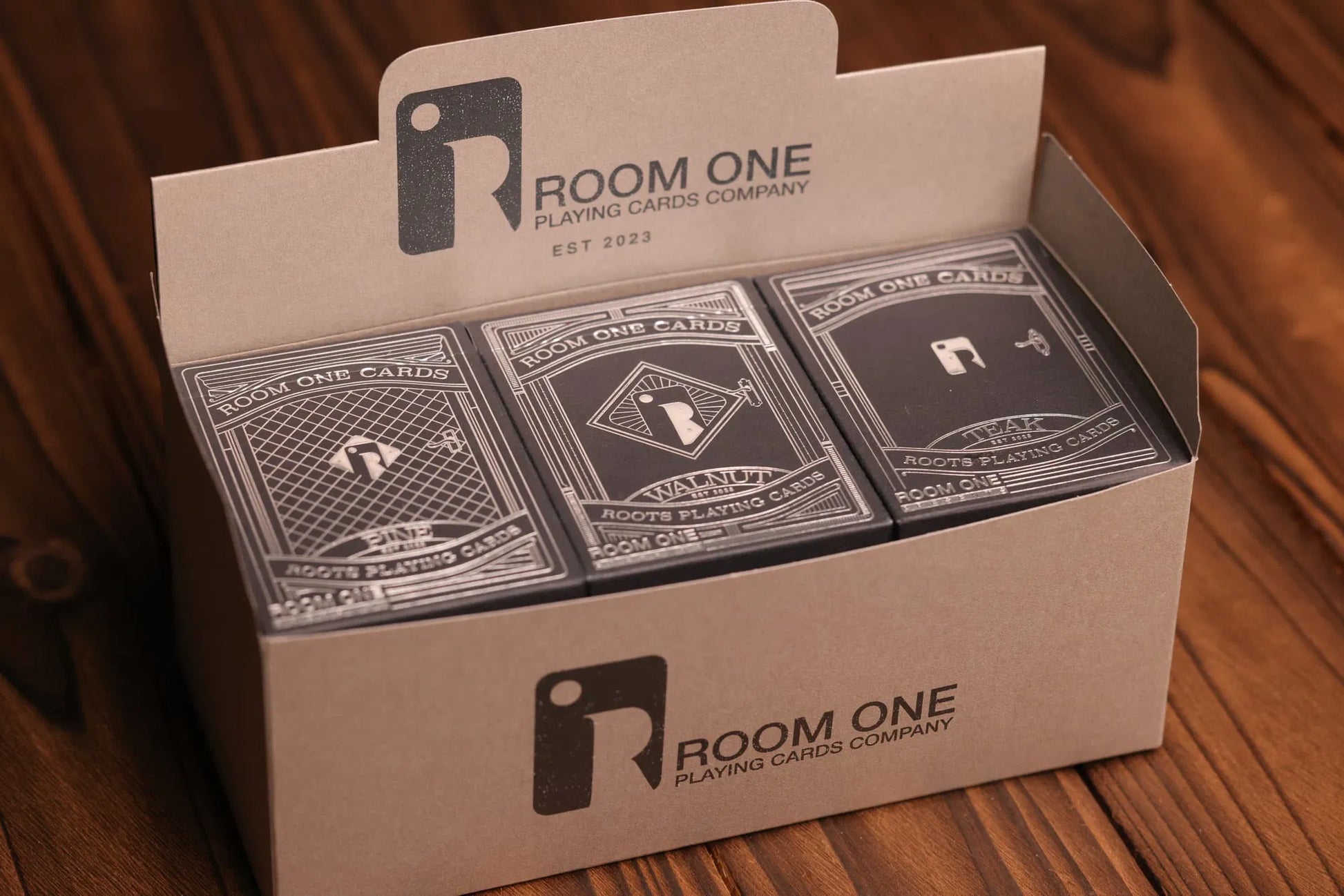 A box containing 12 decks of Roots playing cards in columns of 3, with each column displaying a different deck, placed on a wooden surface.
